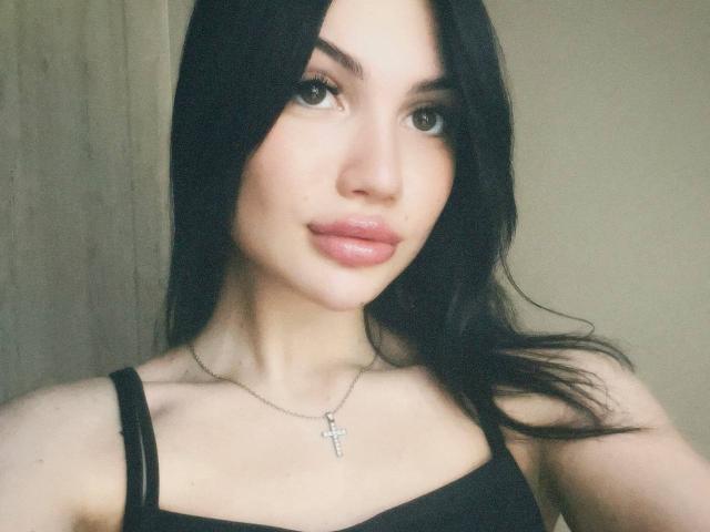 Natalia Liukko a les cheveux noirs foncés, une peau claire et des lèvres roses brillantes. Elle porte un haut noir à bretelles avec un collier en argent en forme de croix. Ses seins sont petits et mignons..