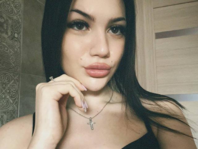 Natalia Liukko mit langem, schwarzem Haar, vollen Lippen und einer Kreuzkette, ihre Hand berührt ihr Kinn, wobei ein kleiner Schönheitsfleck unter ihrer Nase sichtbar ist, sie trägt ein schwarzes Oberteil..