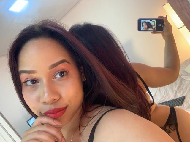 SusanBloom with red lipstick, black bra, and black panties, taking a selfie, showing her back and butt in the mirror, holding a phone, long dark brown hair, smiling.