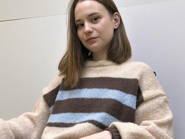 AdelinaGordo with straight brown hair, light skin, and a small nose, wearing a beige and brown striped knit sweater, looking slightly to the side with a subtle smile.