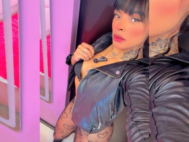 EmmaEbony with black leather jacket, tattoos, fishnet stockings, and pink lipstick, exposing her tits, holding jacket, black bra, purple wall, and mirror reflection, looking up.
