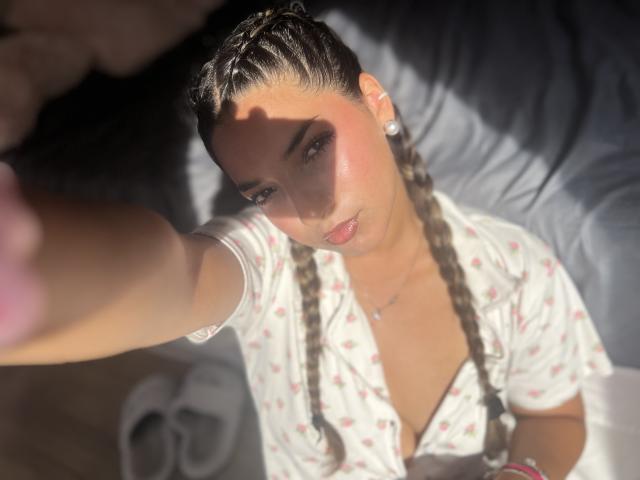 AmbarDunn with braided hair, pink blush, and open shirt, holding out arm, sunlight on face, pearl earrings, looking up, pink lips, white shirt with small red patterns.