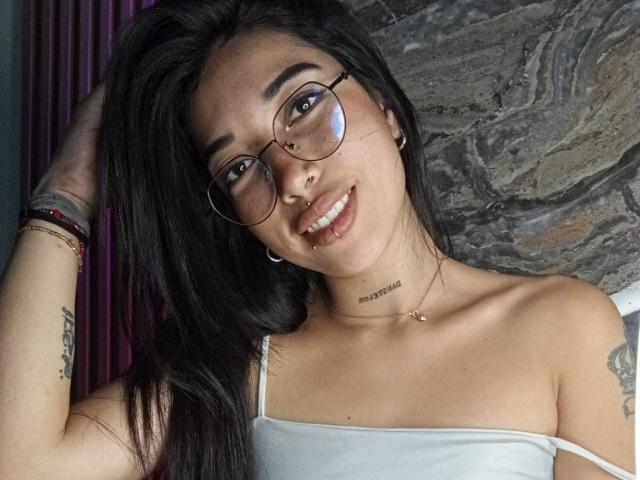 KrystalEvans with long black hair, glasses, smiling, white tank top, nose ring, septum piercing, tattoo on arm, necklace, hand in hair, tattoos on arm.