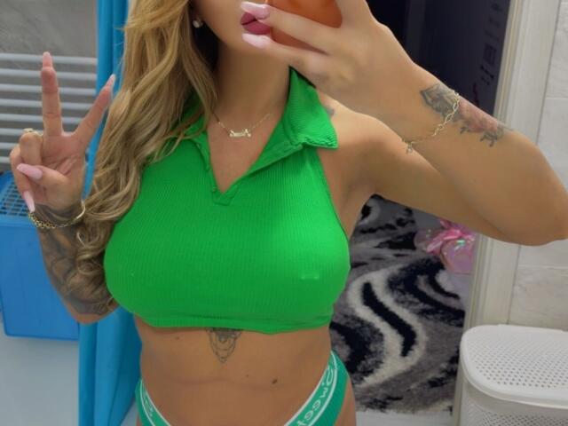 NicolSNaya with green ribbed crop top, showing her big tits, green panties, tattoo below her belly, long blonde hair, pink nails, peace sign, holding phone, wearing gold necklace.