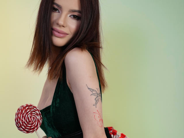 AmirraLovely with brown hair, green dress, holding red swirl candy, butterfly tattoo on right arm, slight smile, pale skin, looking at camera. Boobs visible, cute, sexy.
