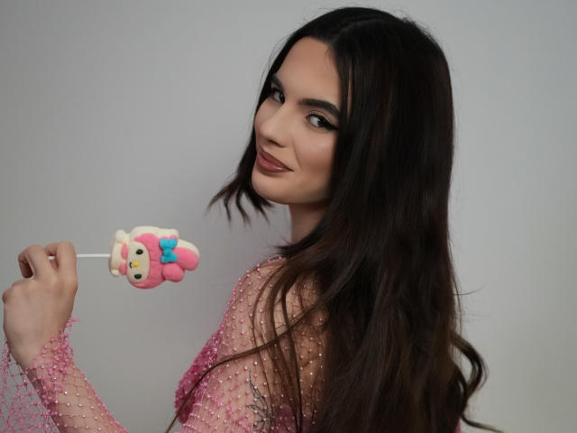 AmirraLovely with long dark hair, pink sparkly dress, holding a cute pink and white cartoon lollipop, smiling at the camera, showing off her tits.