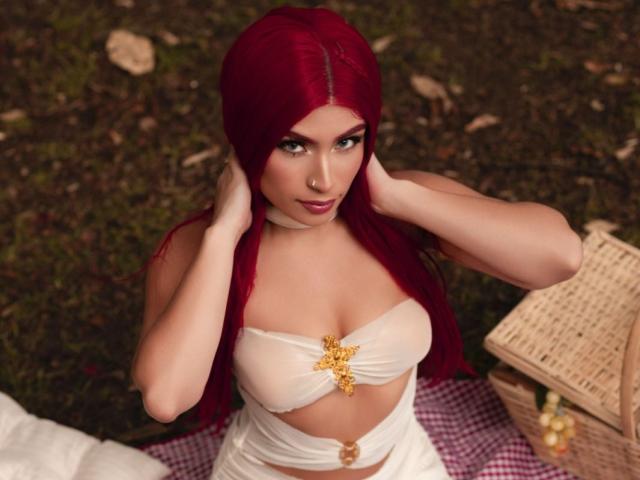 TifanySuarez with red hair, white strapless top, and gold flower, hands in hair, sitting on a red checkered blanket, wicker basket to the right, looking up.