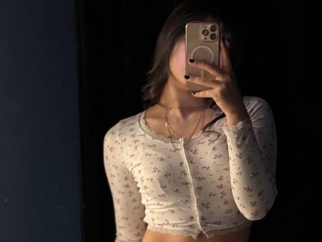 AlidaKarma taking a mirror selfie, wearing a white floral crop top with long sleeves, black nails, holding a gold iPhone, face mostly hidden, dark background.