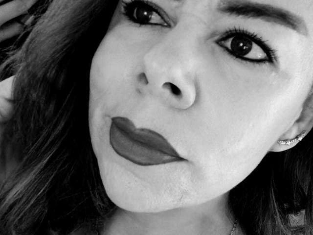 Black and white selfie of EmiliaMilf with thick eyebrows, dark eyeliner, and full lips. She's sticking out her tongue slightly. Her right hand is touching her head.