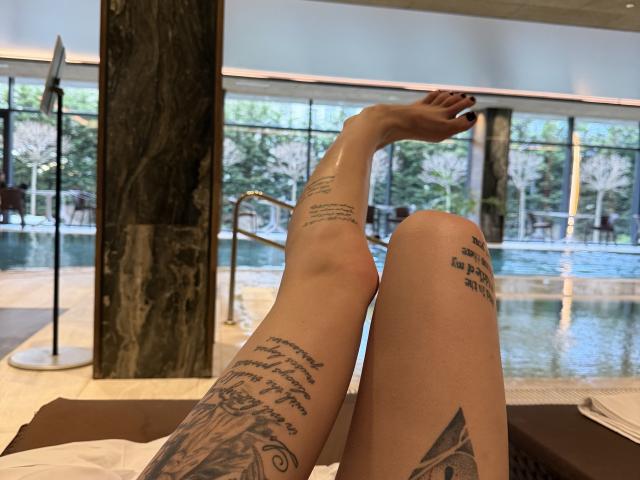 SensualMila's tattooed legs, one foot raised, showing off black ink text and a detailed tattoo on her right calf, near a marble column and a pool.
