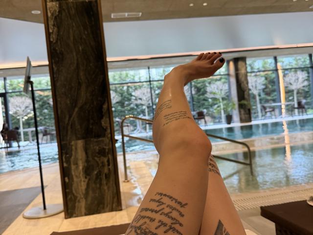 SensualMila's legs are crossed at the ankles, showing off tattoos and painted red toenails, with her feet pointing upwards in a marble-walled indoor pool area.
