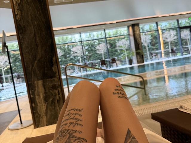 SensualMila's legs are in a pool's indoor area, showing off tattoos on both thighs. Her bare knees point forward, with her white panties visible at the bottom.