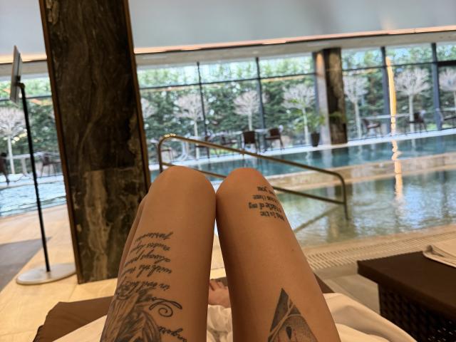 SensualMila's tattooed thighs, showing leg tattoos, in a relaxed indoor pool setting, with her feet up and legs crossed, pool visible in the background.