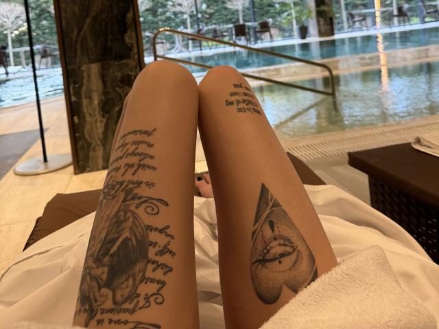 SensualMila's tattooed thighs, lying on a white towel, showing detailed black ink tattoos on both legs, near a pool, with her legs slightly parted.
