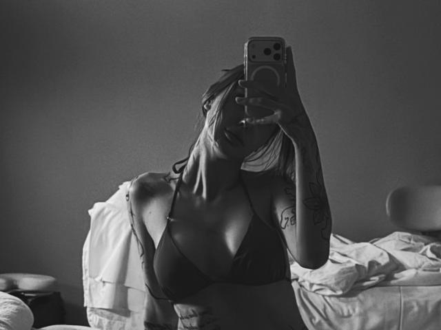 SensualMila with medium tits, dark bra, tattoos, holding phone, mirror selfie, grey background, messy bed, off-shoulder shirt, arm tattoo, phone with ring, blonde hair, monochrome image.