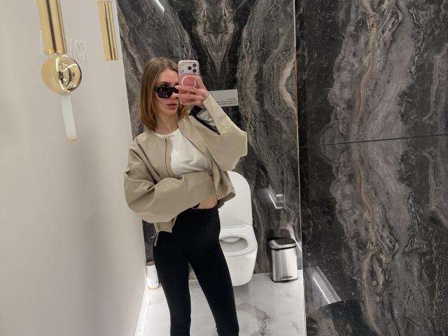SensualMila with blonde hair, black sunglasses, beige jacket, white top, black leggings, holding pink phone, taking mirror selfie in marble bathroom.
