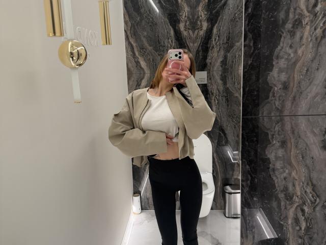 SensualMila with blonde hair, wearing a beige jacket, white crop top, and black pants, taking a mirror selfie in a marble bathroom, showing her tits and ass.