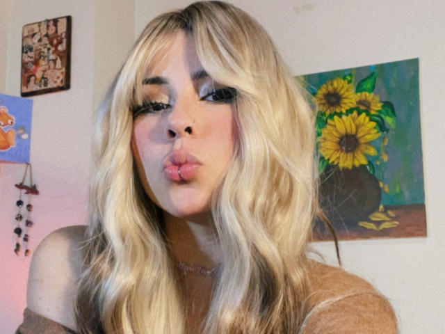 Blond GaiaJoness with pouty lips, wearing a brown top, has wavy hair, thick eyebrows, and is making a kissy face in a selfie. Boobs slightly visible under top.