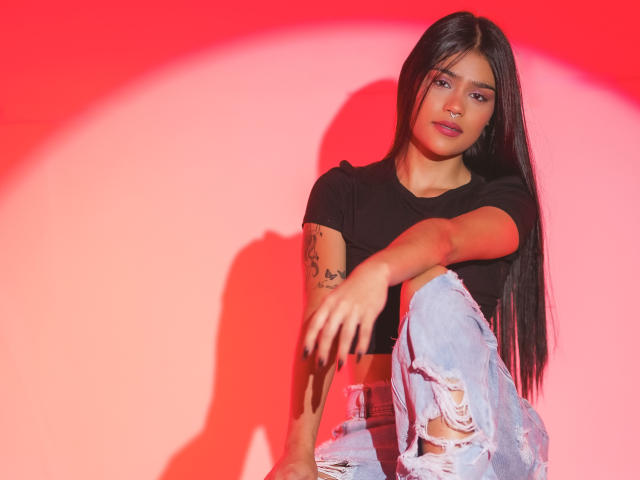 DevourFire with long black hair, wearing a black crop top and ripped jeans, has a tattoo on her arm, sitting with one leg up, red light casting a shadow.