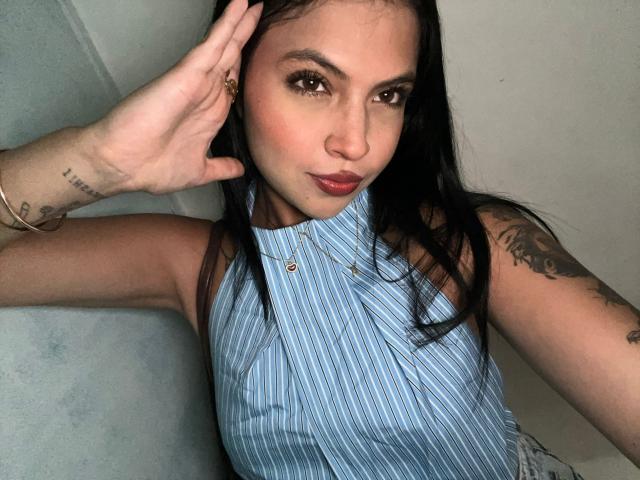 NyraVanel with long black hair, brown eyes, and red lipstick, wearing a blue-striped sleeveless top, showing tattoos on her arm, hand touching her head. NyraVanel's face is centered.