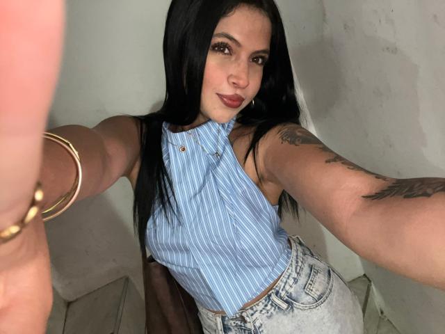 NyraVanel, con cabello largo y negro, vestida con una camiseta azul a rayas sin mangas y jeans de cintura alta, mostrando tatuajes en su brazo derecho y una pulsera dorada, tomándose un selfie..