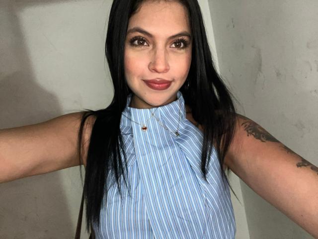 NyraVanel with long black hair, wearing a blue striped sleeveless shirt, arms raised, showing tattoos, and a confident smile, brown eyes, and a nose ring.
