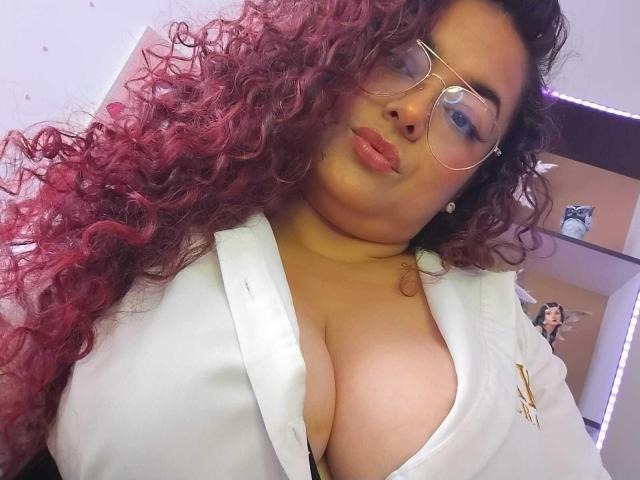 SophiLarusso with curly red hair, wearing glasses, pouty lips, and a white shirt that shows her big, juicy tits. Her chest is prominently featured in the selfie.