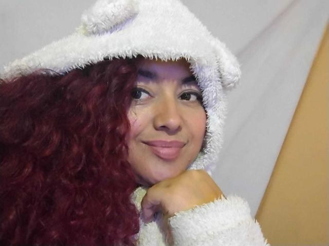 SophiLarusso with curly red hair, wearing a fluffy white animal hood, smiling softly, her hand under her chin, in a casual, cozy pose.