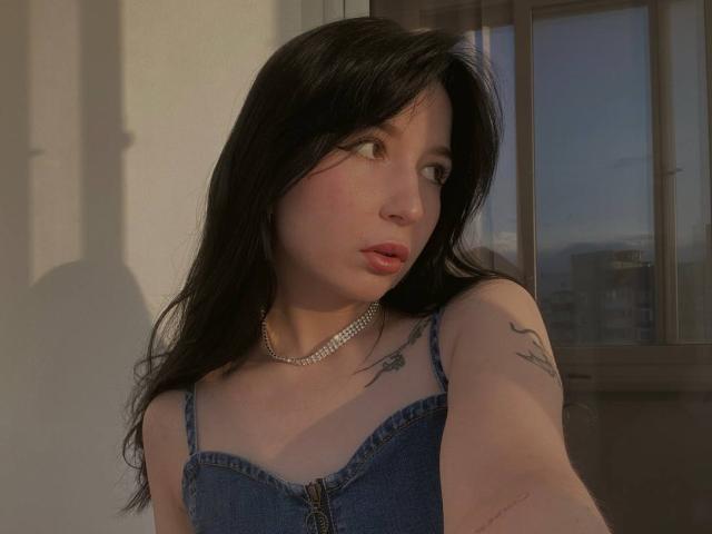 MertieDasinger with long black hair, pink lips, and a denim dress, showing tattoos on her right shoulder, wearing a pearl necklace, taking a selfie.
