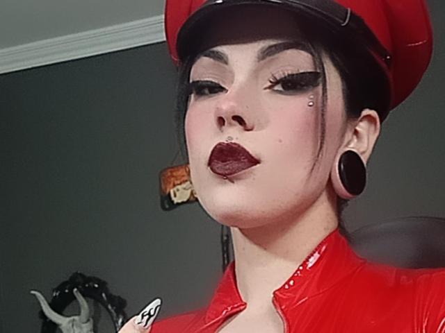 AmyDan69, avec du rouge à lèvres foncé, de l'eye-liner noir et un petit piercing au nez, portant une casquette rouge et une veste en cuir rouge assortie, des élargisseurs d'oreilles noirs et un léger sourire..