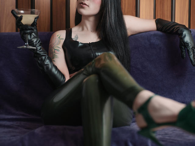 AmyDan69 mit langen, schwarzen Haaren, Lederhandschuhen, einem schwarzen Tanktop, grünen Leggings, die ein Martini-Glas hält, ein Tattoo am linken Arm, mit verschränkten Beinen, grünen High Heels, sitzend auf einem violetten Sofa..