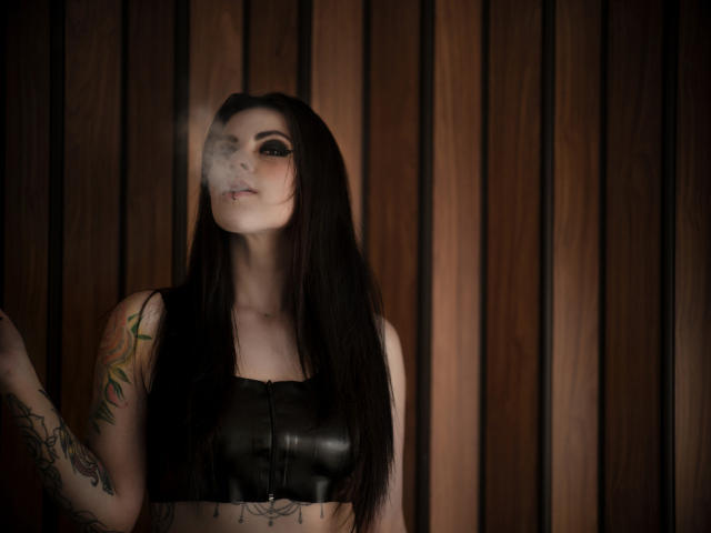 AmyDan69, con lunghi capelli neri, che fuma, indossa un top corto in pelle nera, ha tatuaggi sulle braccia, guarda di lato, sfondo di una parete di legno, atmosfera cupa e malinconica. Seno, capezzoli e glutei non sono visibili..