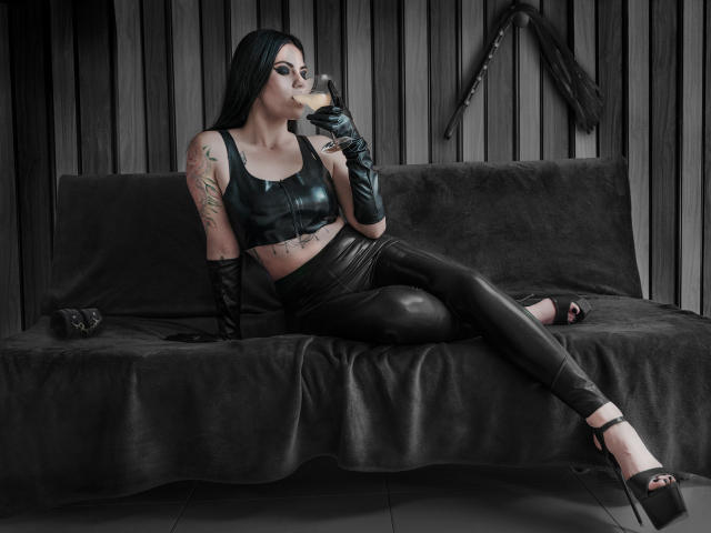 AmyDan69 con un top corto de cuero negro, pantalones negros brillantes, guantes y tacones, fumando, con tatuajes en el brazo, sentada en un sofá negro, tacones altos negros, pared de madera, látigo en el fondo..