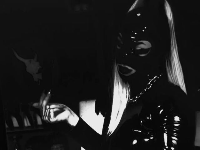 AmyDan69 portant un masque en latex noir avec des yeux blancs, de longs cheveux blonds, vêtue d'une tenue en latex brillante, tenant une chaîne, sur une photo en noir et blanc sombre et atmosphérique..