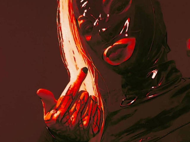 AmyDan69 con una máscara y guantes de látex negro brillante, lápiz labial rojo, levantando un dedo hacia el rostro con una luz roja y naranja intensa. Los senos no son visibles. Las manos y el rostro están resaltados..
