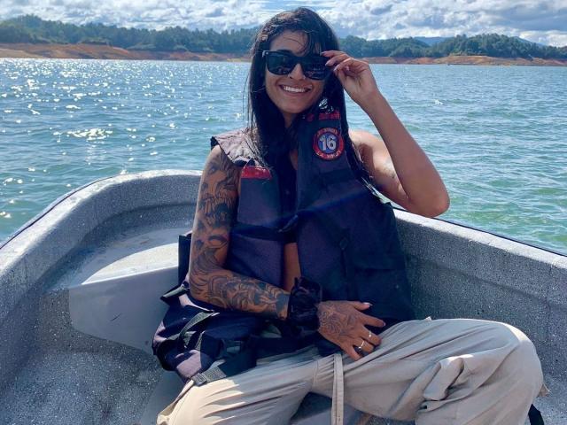 RoxyThunder with tattoos, wearing a black life vest, sunglasses, and beige pants, sitting in a boat, smiling, adjusting her sunglasses, with blue water and a cloudy sky.