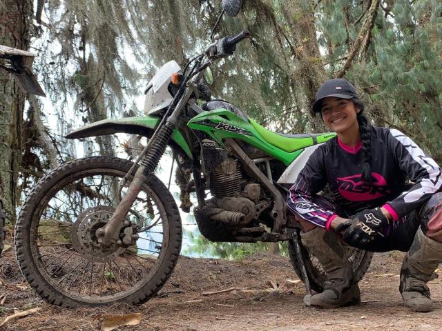 RoxyThunder, crouching next to a muddy green Husqvarna dirt bike, wearing black helmet, gloves, and pink/white motocross gear, smiling with dirty brown pants.