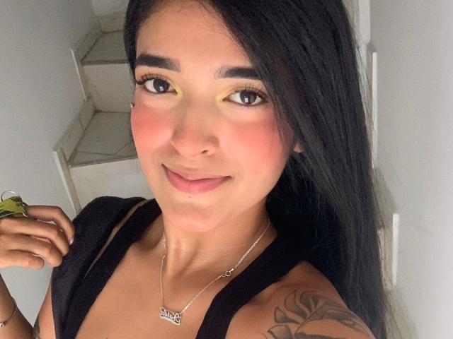 RoxyThunder, avec de longs cheveux noirs, du fard à paupières jaune, du blush rose, un débardeur noir, un collier en argent, un tatouage sur l'épaule droite, tenant une feuille verte, souriant à la caméra..
