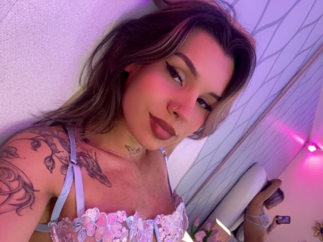LenaBranch, com cabelo castanho claro, delineador rosa e tatuagens no ombro, vestindo um sutiã de renda azul, sorrindo para a câmera. Seus seios são pequenos e ela tem um piercing no nariz..