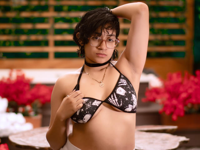 DaryMoon mit kurzen, schwarzen Haaren, Brille, schwarzem Choker und einem schwarz-weißen Bikini-Oberteil mit Blumenmuster, Arm erhoben, Hand auf der Brust, zeigt kleine Brüste, sanfter Gesichtsausdruck, rosa Blumen im Hintergrund..