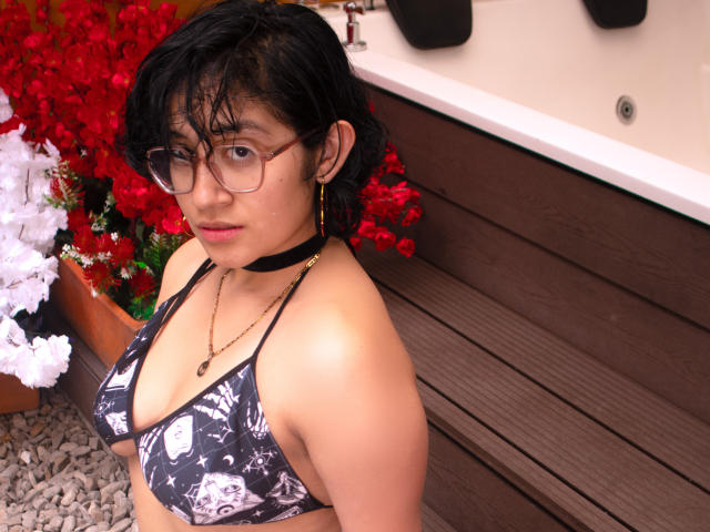 DaryMoon has dark hair, glasses, and a black choker, wearing a patterned bikini top, showing her small boobs and tan skin, with a serious expression, standing by a hot tub.