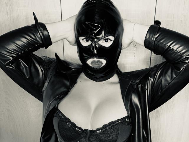 SlaveKaate indossa una maschera di lattice nero, una giacca di pelle e un reggiseno di pizzo, con le braccia alzate, mostrando i suoi seni. Indossa guanti neri e un braccialetto, in una foto in bianco e nero..