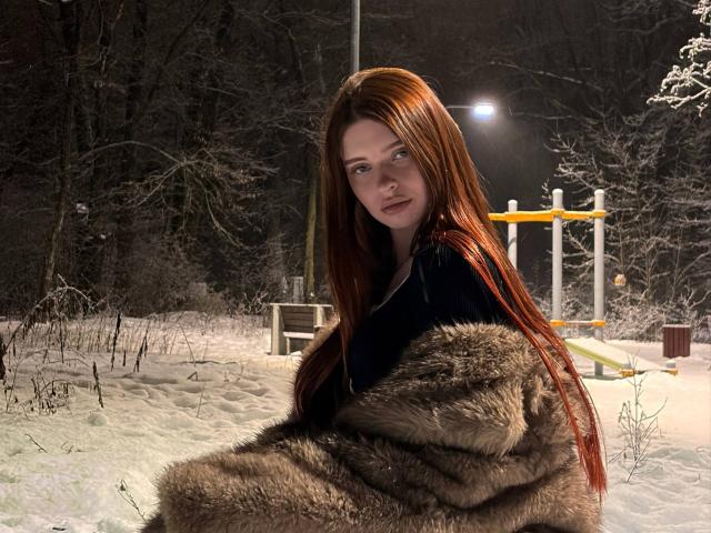 LeidaAtengco with long red hair, wearing a black top and brown fur coat, sitting in snowy forest at night, looking directly at the camera with a serious expression.