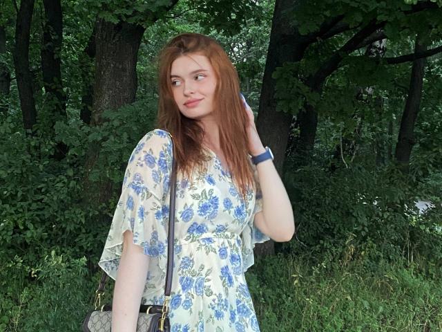 LeidaAtengco with red hair, blue floral dress, smiling, hand in hair, black crossbody bag, green forest background, wearing blue wristwatch, standing on grass, looking to the side.