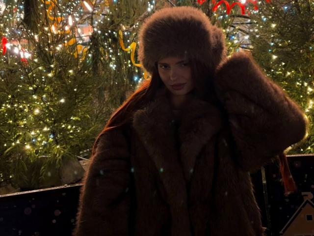 LeidaAtengco in a fluffy brown fur hat and coat, touching her head with a slight smirk, surrounded by Christmas lights and greenery, looking sexy and winter-ready.