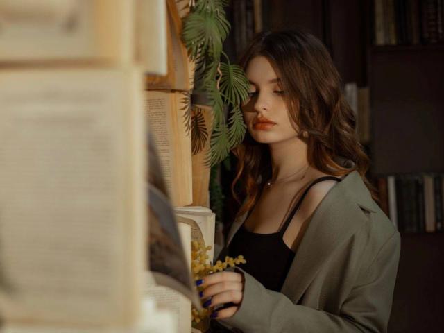 LeidaAtengco with wavy brown hair, wearing a green jacket and black top, reading a newspaper, holding yellow flowers, in a softly lit, cozy room.
