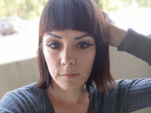 OrianaLaFrancaise has dark brown bob, thick eyebrows, and black eyeliner, wearing a gray sweater, hand in hair, with a small beauty mark below her nose, looking at camera.