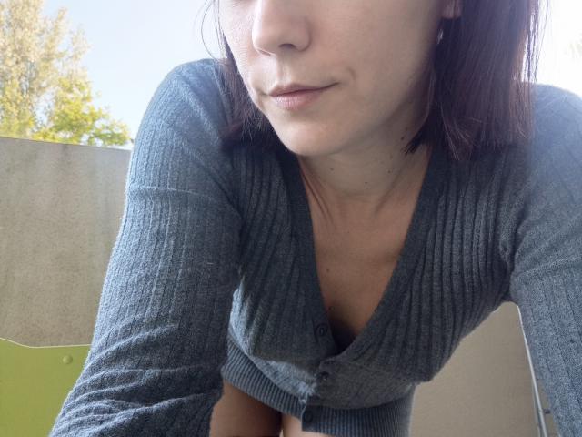 OrianaLaFrancaise, with a slight smirk, wearing a gray, ribbed, V-neck cardigan, showing her small tits, leaning forward, her face partially visible, brown hair, light skin.