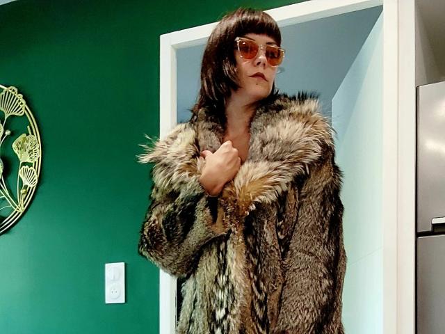 OrianaLaFrancaise with brown bob, wearing glasses and a thick, furry brown coat with a large fur collar, standing in a green-walled room.