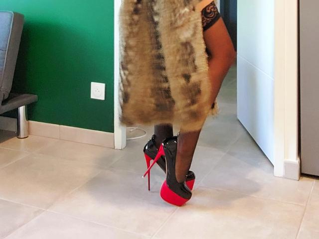 OrianaLaFrancaise's ass in black stockings, wearing a fur coat, and red-and-black high heels, standing on tiled floor, green wall in background.